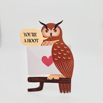 Funny die-cut owl greeting card with the pun “You’re a Hoot.” Great horned owl carrying a folded card, blank for message